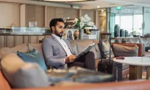 Mann im Anzug liest Zeitschrift in der airport VIP lounge. Sein Koffer steht bereit.