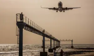 Zwei Personen genießen das Plane Spotting am Strand, während ein Flugzeug über eine Seebrücke hinwegfliegt.