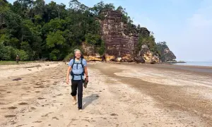 Mann mit Rucksack und Schuhen in der Hand geht barfuß am Strand entlang. Die Packliste für Malaysia ist komplett, die Natur ist üppig.