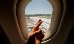 Hand mit überkreuzten Fingern vor Flugzeugfenster. Ein Zeichen der Hoffnung für alle Flugzeug-Passagiere.