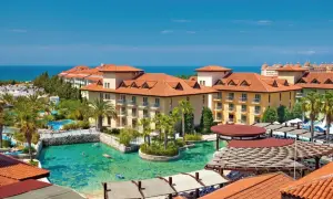 Ein luxuriöser Urlaub im Club Grand Aqua: Sonnige Gebäude mit roten Ziegeldächern, Palmen und ein türkisfarbener Pool mit Badegästen.