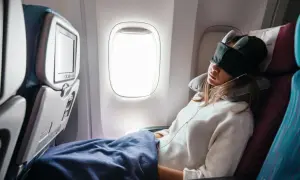 Frau schläft im Flugzeug, mit Schlafmaske und Nackenkissen, eingewickelt in eine Decke.