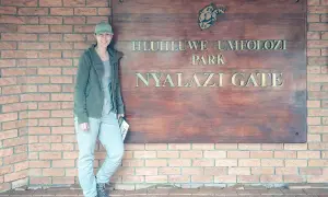 Frau am Eingangstor des Hluhluwe Umfolozi Parks, bereit für ihr Safari-Abenteuer. Ihre Kleidung ist perfekt für die Packliste.