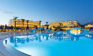 Ein beleuchteter Pool spiegelt das Lindos Princess Beach Hotel wider. Palmen und blaue Sonnenschirme säumen den Poolbereich.