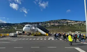 Passagiere steigen am Madeira Flughafen aus dem Flugzeug. Im Hintergrund erstreckt sich eine hügelige Landschaft mit Häusern.