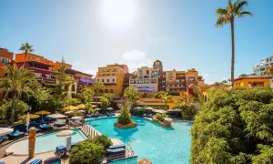 Sonnige Poolanlage mit bunten Gebäuden und Palmen im Riu Palace Teneriffa. Entspannung pur unter blauem Himmel.