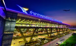 Der beleuchtete Flughafen Bangkok Suvarnabhumi bei Nacht, mit Autos auf mehrstöckigen Zufahrtsstraßen und einem Flugzeug am Himmel.