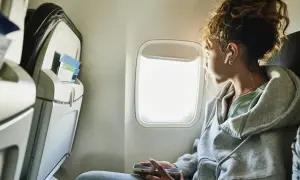 Junge Frau blickt aus dem Flugzeugfenster, vertieft in ihr Handy. Sie fragt sich, wie ist es zu fliegen, während sie die Wolken vorbeiziehen sieht.