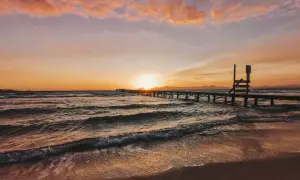 Sonnenuntergang über dem Meer an der Playa de Muro. Wellen brechen sanft am Strand, ein langer Holzsteg führt ins Wasser.