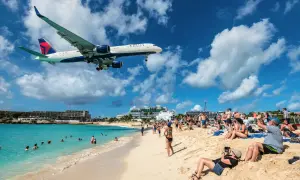Ein Delta-Flugzeug landet tief über einem belebten Strand in der Karibik. Urlauber am **Karibik Flughafen** genießen die Sonne, während das Flugzeug über ihnen kreist.