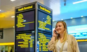 Frau telefoniert am London Flughafen vor einer Anzeigetafel für den South Terminal.