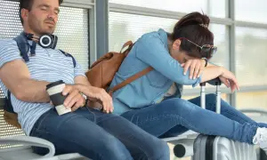 Erschöpftes Paar wartet auf den Flug. Der Mann schläft mit Kaffee, die Frau hat den Kopf auf den Koffer gelegt. Der Jetlag geht nicht weg.