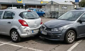 Zwei Autos sind an einem Unfall auf einem Parkplatz beteiligt. Der silberne Opel hat einen stark beschädigten Stoßfänger, der graue VW hat vorne eine Delle.