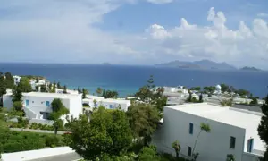Blick auf das Robinson Daidalos Resort mit weißen Gebäuden und blauen Fenstern, umgeben von grüner Vegetation, am Meer mit Inseln am Horizont.
