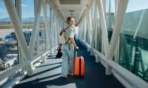 Junge Frau mit orangefarbenem Koffer und Rucksack geht durch einen Flugsteig. Ihr Flug mit Zwischenstopp beginnt.