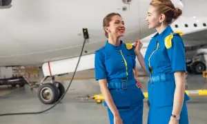 Zwei Flugbegleiterinnen in blauen Uniformen mit gelben Akzenten stehen vor einem Flugzeug und unterhalten sich.