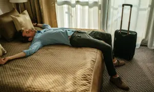Erschöpfter Mann liegt auf einem Hotelbett, Koffer daneben. Der Jetlag von Asien nach Deutschland hat ihn völlig ausgeknockt.