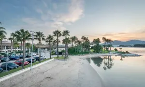 Strand mit Palmen und dem Hotel Alcudiamar im Hintergrund, ruhiges Wasser spiegelt den Himmel.