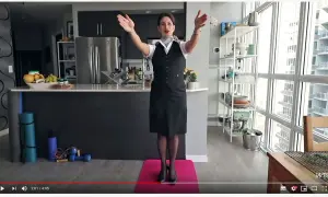 Eine Stewardess oder Flugbegleiterin in Uniform steht auf einer rosa Matte und macht Übungen. Im Hintergrund eine Küche und ein großes Fenster.