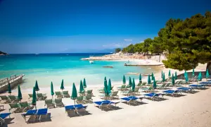 Ein sonniger **Tirana Strand** mit türkisfarbenem Wasser, Liegestühlen und Sonnenschirmen.