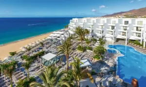 Traumhafter Blick auf das Riu Palace Jandia: Weißes Hotel, Pool, Palmen und der goldene Strand am türkisfarbenen Meer.