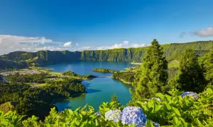 Grüne Hügel umrahmen einen tiefblauen See. Hier liegen die Azoren, eine Inselgruppe im Atlantik, mit üppiger Vegetation und blühenden Hortensien im Vordergrund.