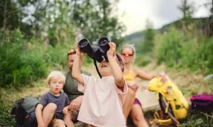 Familie im Urlaub mit Kleinkind: Ein Mädchen schaut durch ein Fernglas, während ihr Bruder und die Eltern im Hintergrund sitzen.