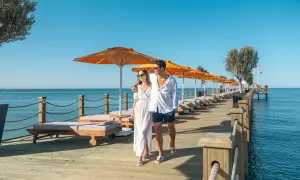 Ein Paar spaziert über einen Holzsteg am Meer, gesäumt von Liegen und Sonnenschirmen. Ein perfekter Urlaub im Arum Barut Collection.