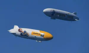 Zwei Zeppeline, einer mit einem Bagger und "Zeppelin CAT", der andere mit "Europa Park" und einem Koala, schweben am blauen Himmel.