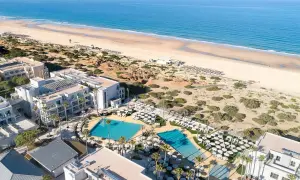 Luftaufnahme des Hipotels Barrosa Park mit Pools, Liegestühlen und Palmen, daneben ein breiter Sandstrand und das blaue Meer.