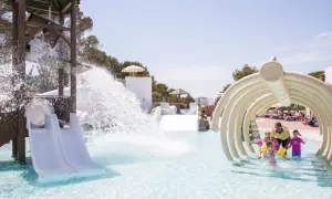 Kinder planschen im Wasserpark, während ein Wasserfall vom Rutschenturm herabstürzt. Ein Mann mit gelbem Shirt beaufsichtigt die Kleinen unter einem knochenartigen Tunnel in den Calimera Fido Gardens.