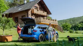 Familie packt für den Urlaub mit E-Auto. Das blaue Elektroauto wird vor einem Holzhaus geladen, während der Hund im Garten wartet.