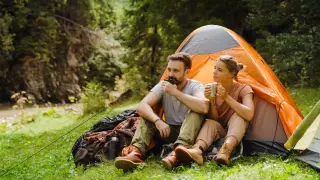Ein Paar genießt die Ruhe beim Zelten gehen. Sie sitzen vor ihrem orangefarbenen Zelt, trinken aus Tassen und blicken in die grüne Natur.