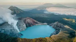Der Ijen-Krater auf der **Java Insel** mit seinem türkisfarbenen Säuresee und rauchenden Vulkanflanken, umgeben von grünen Bergen und Wolken.