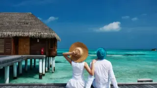 Ein Paar genießt die Aussicht auf das türkisfarbene Meer von einer Holzterrasse. Sie sitzen nebeneinander, die Frau mit einem großen Strohhut, der Mann mit einem blauen Hut. Dies ist, was man in den Flitterwochen macht: entspannen und die Zweisamkeit g...