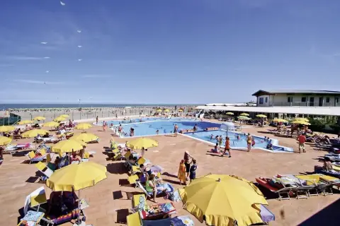 Sonnige Poolanlage mit vielen Liegen und gelben Sonnenschirmen am Rosapineta Sud. Kinder planschen im Wasser, während Möwen über dem Meer kreisen.