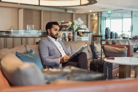 Mann im Anzug liest Zeitschrift in der airport VIP lounge. Sein Koffer steht bereit.