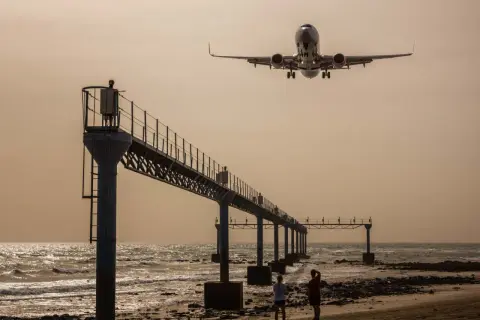 Zwei Personen genießen das Plane Spotting am Strand, während ein Flugzeug über eine Seebrücke hinwegfliegt.