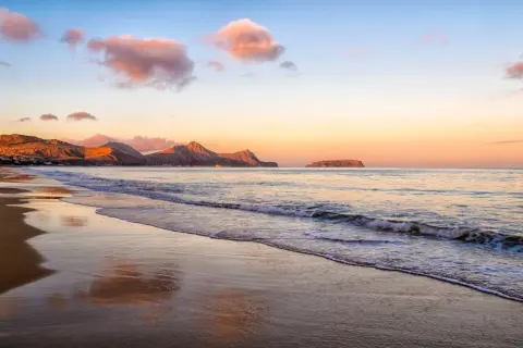 Sonnenuntergang am Strand mit sanften Wellen. Ein idyllischer badeurlaub im winter, mit Bergen im Hintergrund und rosa Wolken am Himmel.