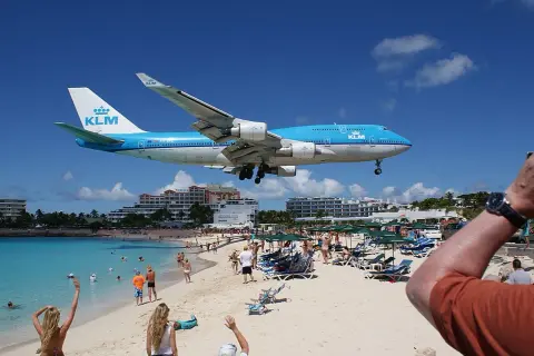 Ein KLM-Flugzeug landet tief über dem Maho Beach Flughafen. Urlauber am Strand winken dem Jumbo-Jet zu.