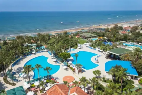 Blauer Poolbereich mit Palmen und Liegen, daneben ein langer Sandstrand mit vielen Sonnenschirmen und dem Meer. Ein Paradies am Strand.