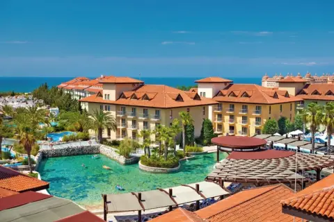 Ein luxuriöser Urlaub im Club Grand Aqua: Sonnige Gebäude mit roten Ziegeldächern, Palmen und ein türkisfarbener Pool mit Badegästen.