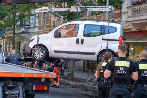 Polizei schleppt ein weißes Auto ab. Ein Mann in orangefarbener Arbeitskleidung hilft. Die auto abschleppen kosten sind hier wohl nicht das Thema.