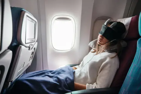 Frau schläft im Flugzeug, mit Schlafmaske und Nackenkissen, eingewickelt in eine Decke.