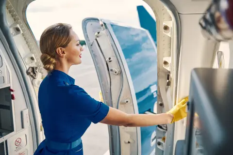 Eine Stewardess in blauer Uniform und gelben Handschuhen reinigt den Eingangsbereich eines Flugzeugs. Ihr Job erfordert Sorgfalt und Sauberkeit.
