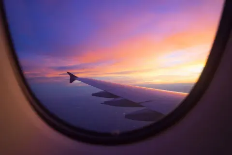 Flugzeugfenster mit Flügel und einem atemberaubenden Sonnenuntergang in Violett- und Orangetönen. Perfekt, um einen **stopover flug buchen** zu planen.