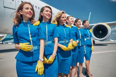 Fünf Flugbegleiterinnen in blauen Uniformen und gelben Handschuhen stehen vor einem Flugzeug. Sie sind bereit, ihre Passagiere zu begrüßen, denn das ist, was man als Flugbegleiterin macht.
