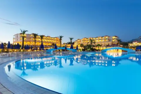 Ein beleuchteter Pool spiegelt das Lindos Princess Beach Hotel wider. Palmen und blaue Sonnenschirme säumen den Poolbereich.