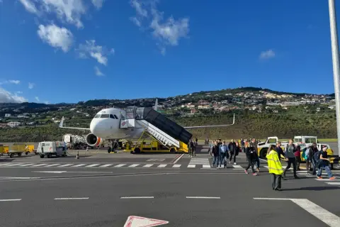 Passagiere steigen am Madeira Flughafen aus dem Flugzeug. Im Hintergrund erstreckt sich eine hügelige Landschaft mit Häusern.
