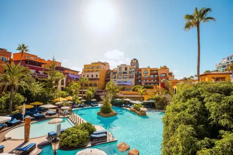 Sonnige Poolanlage mit bunten Gebäuden und Palmen im Riu Palace Teneriffa. Entspannung pur unter blauem Himmel.